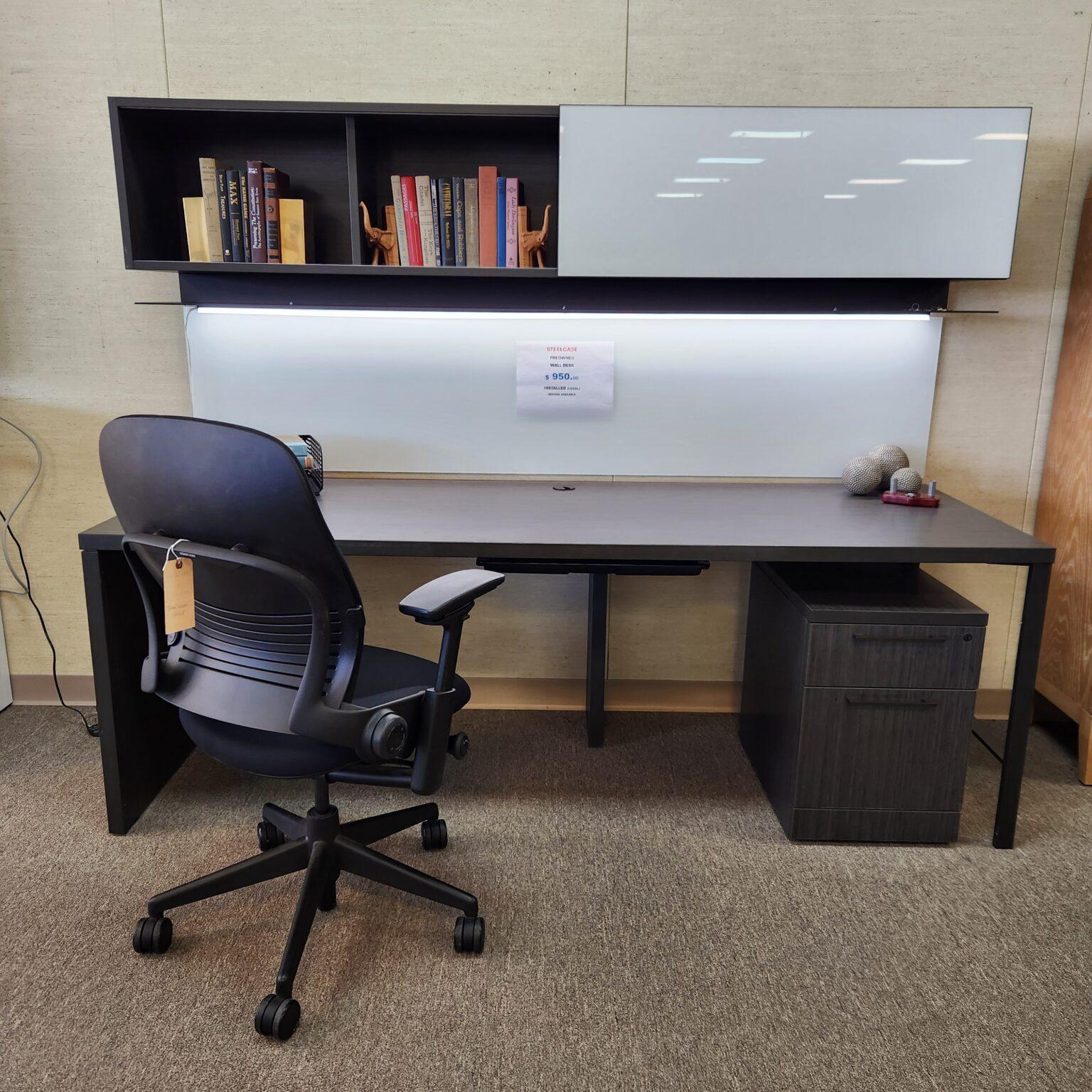 A black Steelcase Leap ergonomic office chair is positioned at a dark gray rectangular desk in a showroom setting. Above the desk, a wall-mounted hutch features open shelving with books and a sliding white glass door. A white whiteboard or privacy panel is mounted between the desk and the hutch, featuring a sign identifying the setup as a preowned Steelcase Wall Desk priced at $950.00. The desk includes a matching dark wood-grain two-drawer mobile pedestal tucked underneath on the right side.