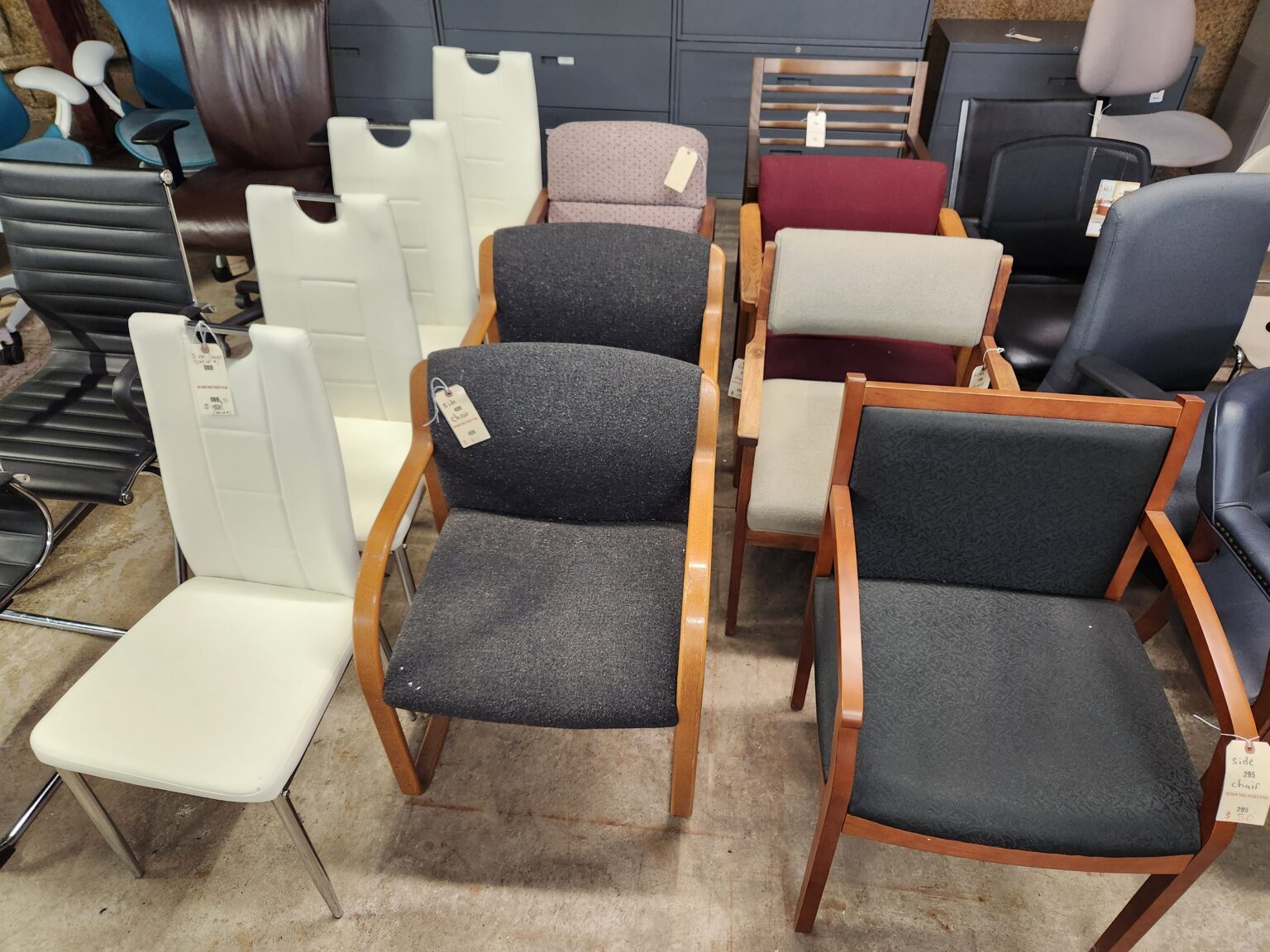A collection of assorted office and side chairs are arranged in rows on a concrete floor in a warehouse or showroom setting. The variety includes modern white leather dining-style chairs with chrome legs, traditional wooden-framed armchairs with dark fabric upholstery, and several ergonomic office desk chairs in black and navy blue. Many of the chairs have paper price tags attached to their frames or armrests, and large gray metal filing cabinets are visible against the wall in the background.