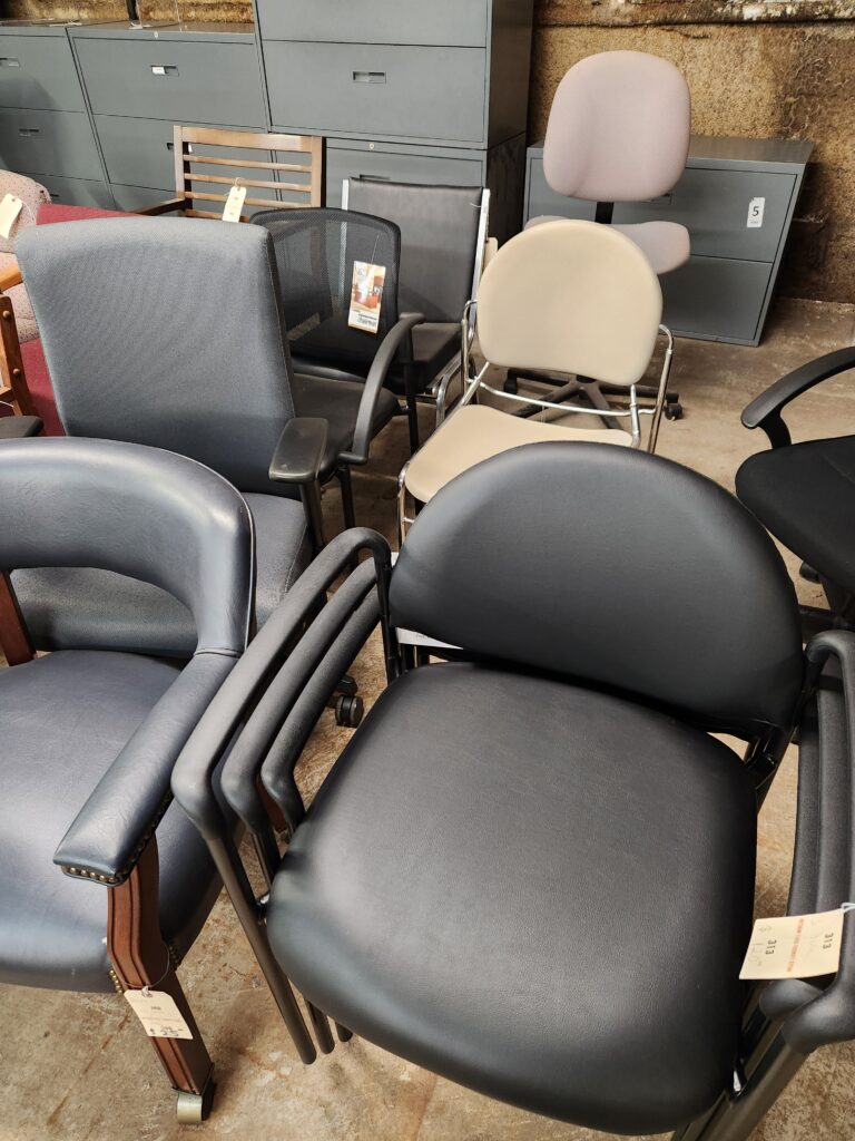 Rows of various secondhand office and guest chairs are arranged in a warehouse setting on a concrete floor. The collection includes white leather-style high-back chairs, dark gray and burgundy fabric armchairs with wooden frames, and black mesh and navy blue ergonomic task chairs. Many chairs have paper price tags attached to their arms or frames. Large gray metal filing cabinets are visible against a textured wall in the background.