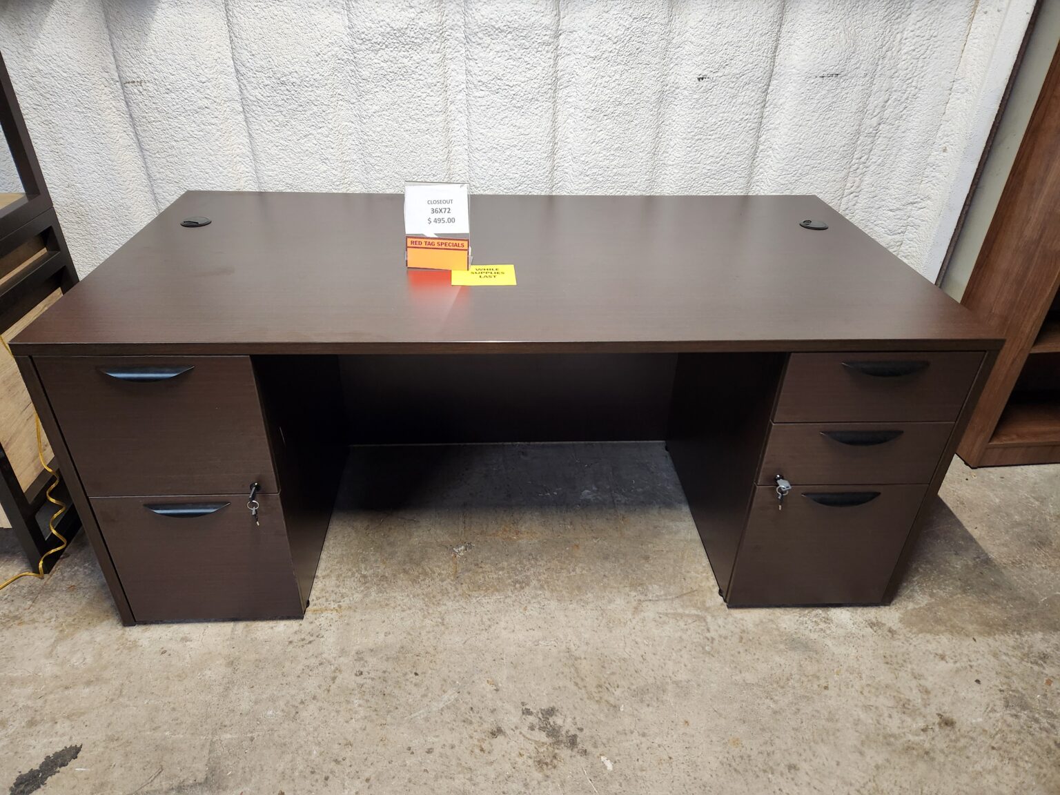 A traditional executive office desk is displayed on a concrete floor in a warehouse setting. The desk features a dark espresso laminate finish and two integrated pedestals: one on the left with two large file drawers and one on the right with three utility drawers. All drawers are equipped with curved, black contemporary handles and locks, with keys inserted into the middle drawer on the right and the top drawer on the left. Two black circular cable management grommets are positioned at the rear corners of the large, rectangular desktop. A clear acrylic stand on the center of the desk holds a "Closeout" sign displaying a price of $495.00 and dimensions of 36x72, with a smaller yellow "While Supplies Last" tag placed next to it.