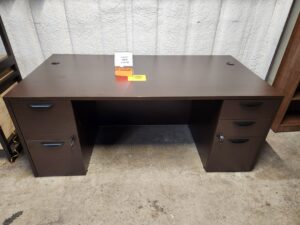 A traditional executive office desk is displayed on a concrete floor in a warehouse setting. The desk features a dark espresso laminate finish and two integrated pedestals: one on the left with two large file drawers and one on the right with three utility drawers. All drawers are equipped with curved, black contemporary handles and locks, with keys inserted into the middle drawer on the right and the top drawer on the left. Two black circular cable management grommets are positioned at the rear corners of the large, rectangular desktop. A clear acrylic stand on the center of the desk holds a "Closeout" sign displaying a price of $495.00 and dimensions of 36x72, with a smaller yellow "While Supplies Last" tag placed next to it.