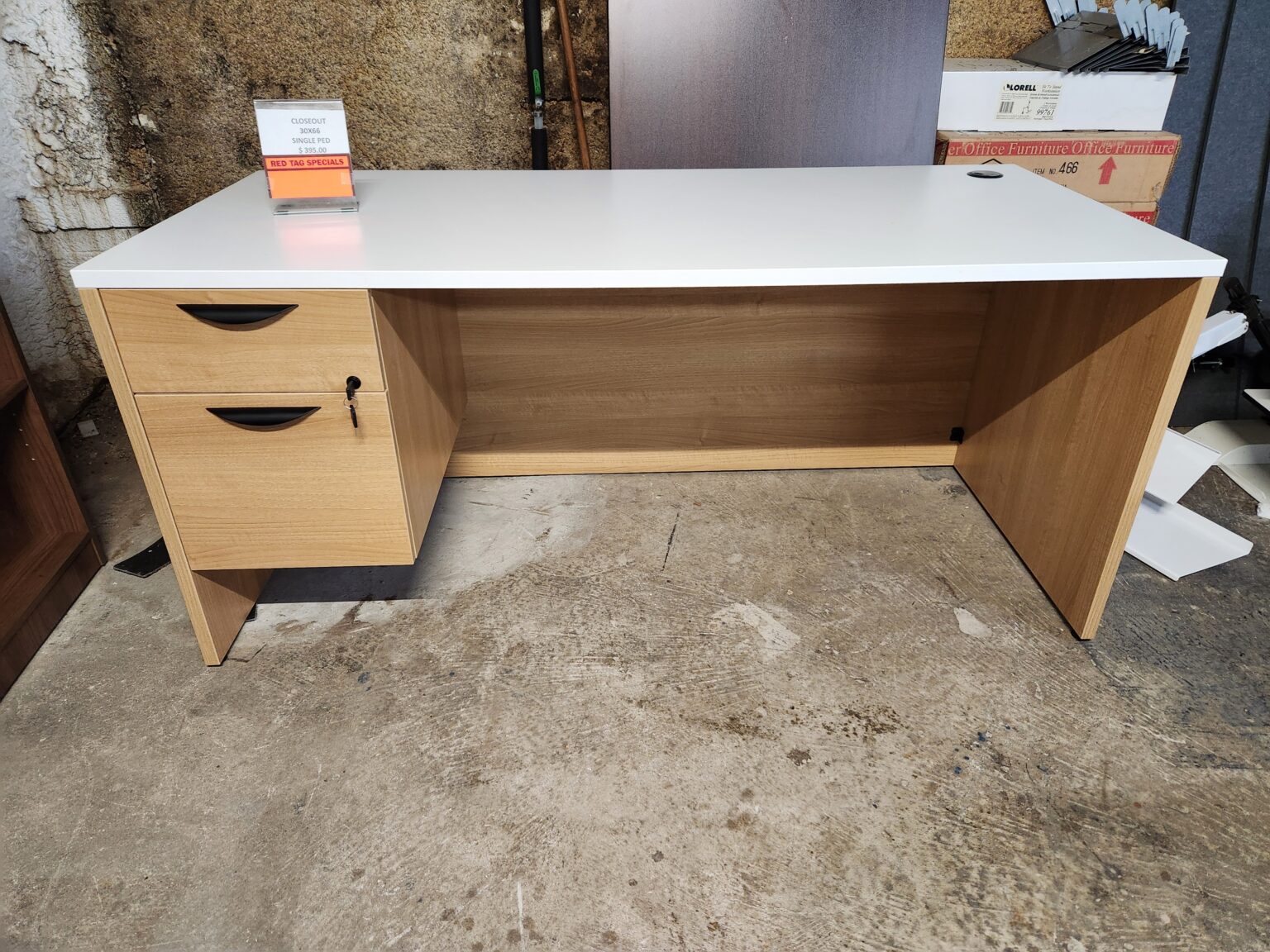 A single-pedestal office desk with a white rectangular desktop and a light oak-finished laminate base is displayed in a warehouse or showroom. The unit features two drawers on the left side: a top utility drawer and a larger bottom file drawer, both with curved black handles and a lock with keys inserted. A clear acrylic sign on the desk indicates a closeout price of $285.00 for the 30x66 single pedestal desk. The background includes partially visible furniture boxes and a textured wall.