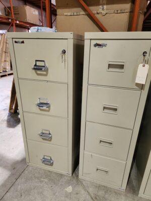 Two beige, 4-drawer vertical fireproof file cabinets are shown side-by-side in a warehouse setting. The cabinet on the left is a FireKing Classic model featuring protruding chrome handles and a key inserted into the top lock. The cabinet on the right is a FireKing Turtle model with recessed drawer pulls and a paper identification tag hanging from the top lock. Both units are designed for space-saving document protection, offering UL-certified fire and impact resistance.