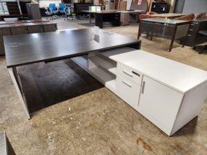 An L-shaped desk workstation is displayed in a warehouse setting, featuring a dark wood-grain rectangular desktop and white support legs. Attached to the side of the main desk is a long white credenza unit that includes two utility drawers with silver horizontal handles and a large storage cabinet. The credenza also features open shelving for additional storage. Other office furniture, including task chairs and filing cabinets, is visible in the blurred background.