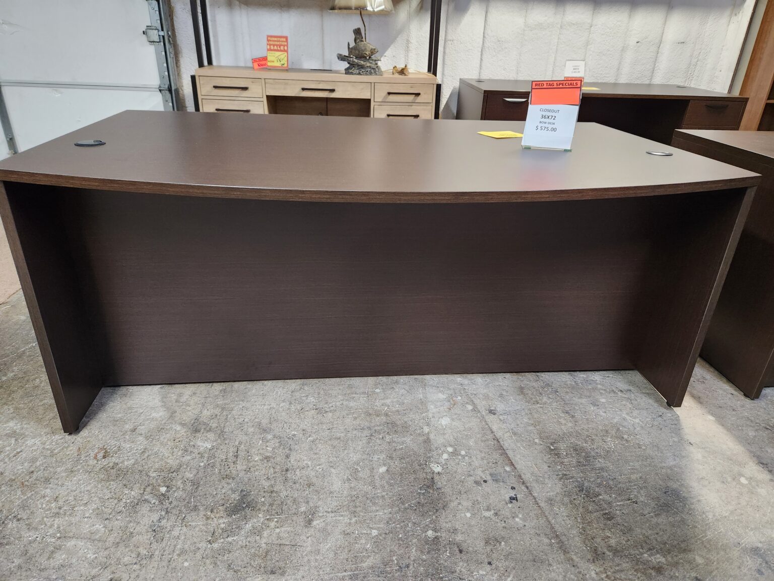 A dark espresso-colored bow-front executive desk is displayed on a concrete floor in a warehouse setting. The large, 36x72-inch rectangular desktop features a slight outward curve along the front edge and two circular black cable management grommets at the rear corners. A clear acrylic sign on the desk indicates a "Closeout" price of $575.00. The desk is a shell-style model with a full-width modesty panel and no integrated storage pedestals. In the background, other office furniture is visible, including a light-colored wood desk with drawers and a decorative lamp.