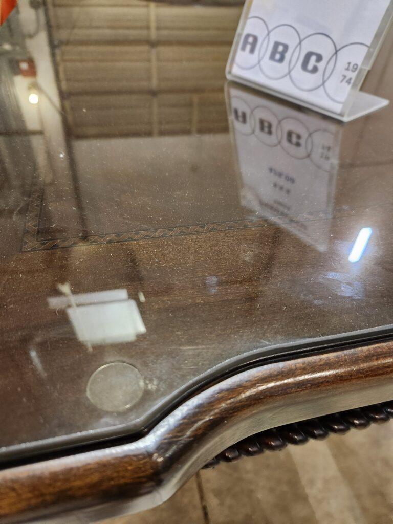 A close-up view shows the corner of a dark wood conference table covered by a protective glass top. The edge of the table is rounded and sits above a carvedtrim that runs along the side of the frame. In the background, a clear acrylic sign with black lettering is partially visible on the table surface.