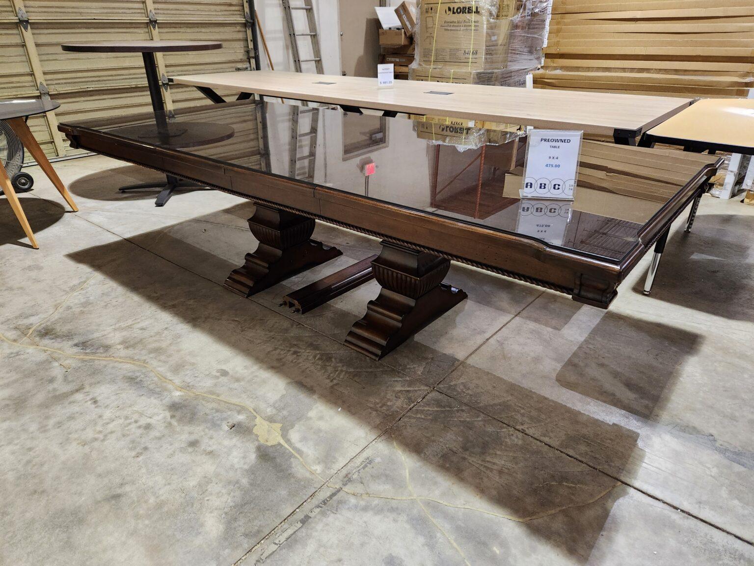 A large, rectangular dark wood conference table is shown from a side angle in a warehouse. The table features a reflective glass top that reveals a decorative wood border. Along the apron of the table is a continuous carved, rope-style trim. It is supported by two massive, ornate pedestal bases with fluted details and tiered, scalloped feet. A clear acrylic sign on the table identifies it as "Preowned" with a price of 475.00.