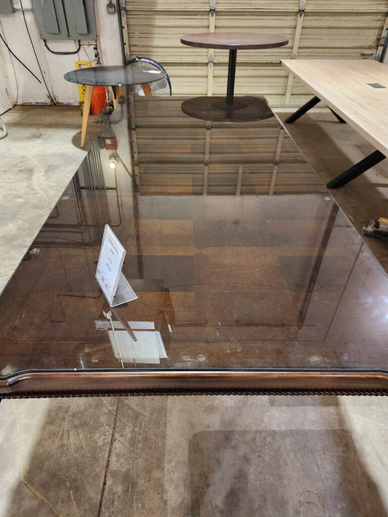 A top-down view of a large, rectangular dark wood conference table covered by a reflective glass top in a warehouse setting. The glass surface shows a decorative wood inlay with a repeating square pattern across the center of the table. A clear acrylic sign indicating the table is preowned and priced at 475.00 sits on the left side of the tabletop. In the background, other office furniture including round and rectangular tables are visible against a warehouse roll up door and concrete floor.
