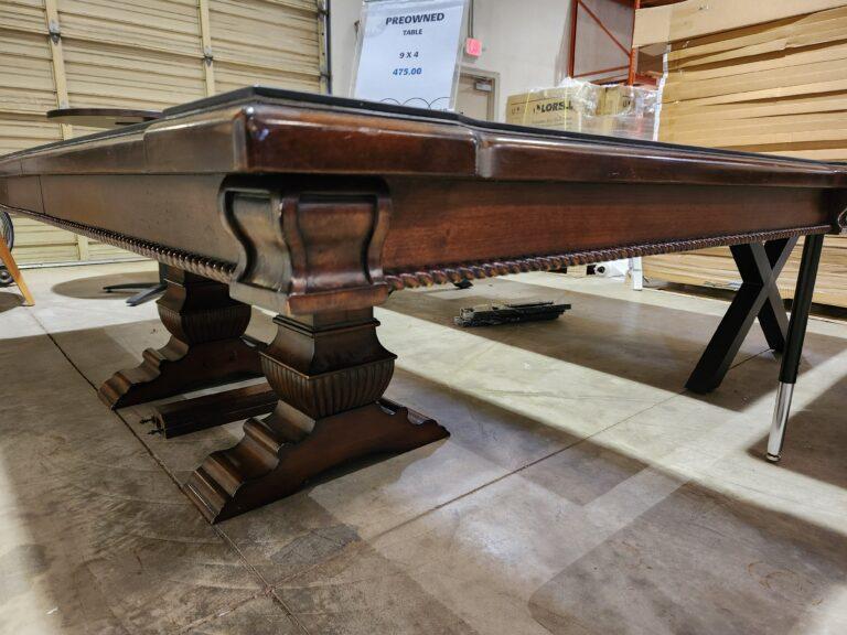 A close-up side profile of a preowned 9-foot by 4-foot rectangular wood conference table displayed in a warehouse setting. The table features a dark, polished wood finish with a thick, decorative edge and a carved, rope-style trim running along the apron. It is supported by massive, ornate pedestal legs with a tiered, scalloped design. A clear acrylic sign resting on the table surface indicates a price of $475.00.