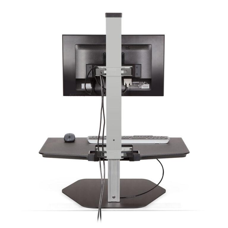 Against a white background, a gray Winston Workstation sit-stand converter is shown with a monitor bracket, keyboard, and mouse. It consists of a large black work surface and a central support pillar with an adjustable monitor mount. The unit sits on a flat, hexagonal metal base and has a small utility tray at the bottom of the support column.