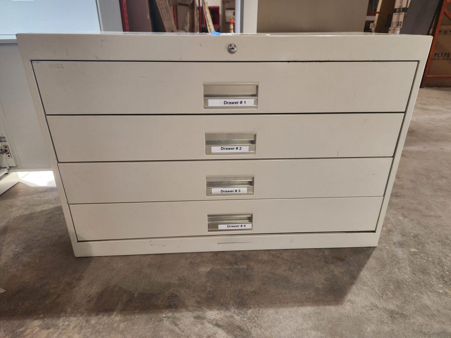 A light gray metal 4-drawer flat file cabinet with a top lock and labeled drawer handles reading "Drawer #1" through "Drawer #4" sitting on a concrete floor in a warehouse setting.
