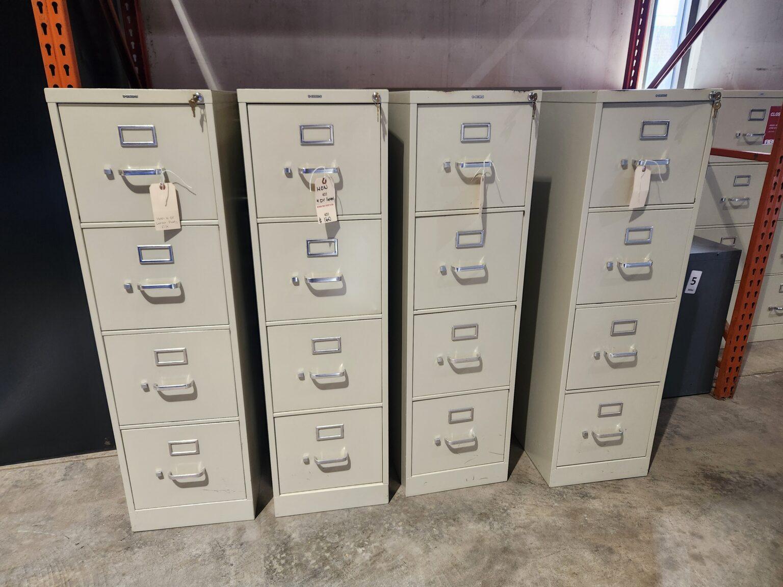 Four beige HON 4-drawer vertical locking letter file cabinets with silver handles and label holders arranged in a row on a concrete warehouse floor.