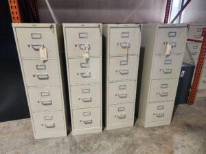 Four beige HON 4-drawer vertical locking letter file cabinets with silver handles and label holders arranged in a row on a concrete warehouse floor.