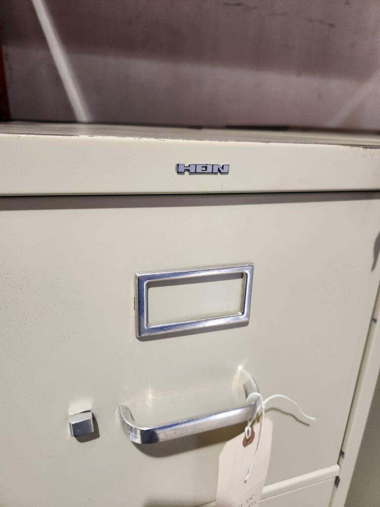 HON; a close-up of a beige metal file cabinet featuring the HON brand logo, a silver label holder, and a silver drawer pull with a paper tag attached.