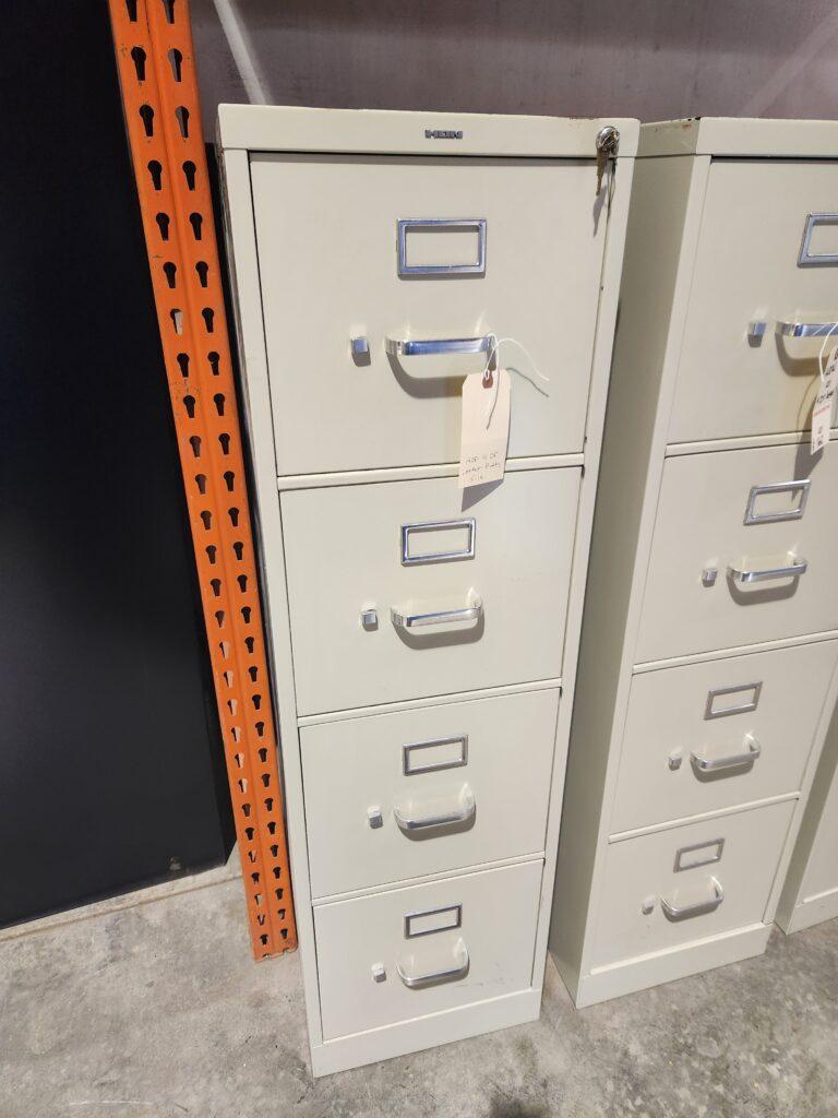 A beige HON 4-drawer vertical locking letter file cabinet with silver handles, label holders, and keys in the top lock, standing in a warehouse next to an orange shelving upright.