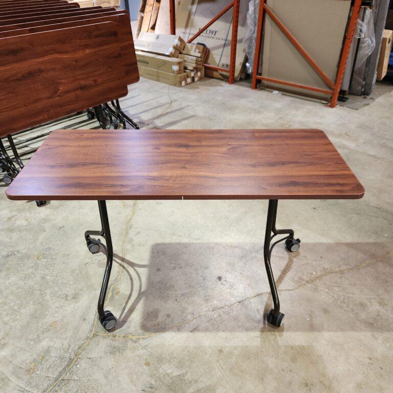 A rectangular training table with a wood-grain laminate top, a dark metal C-leg base, and four casters on a concrete warehouse floor.