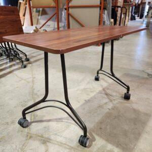 A side-angle view of a rectangular training table with a wood-grain laminate top, a dark metal arched-leg base, and four black casters on a concrete warehouse floor.