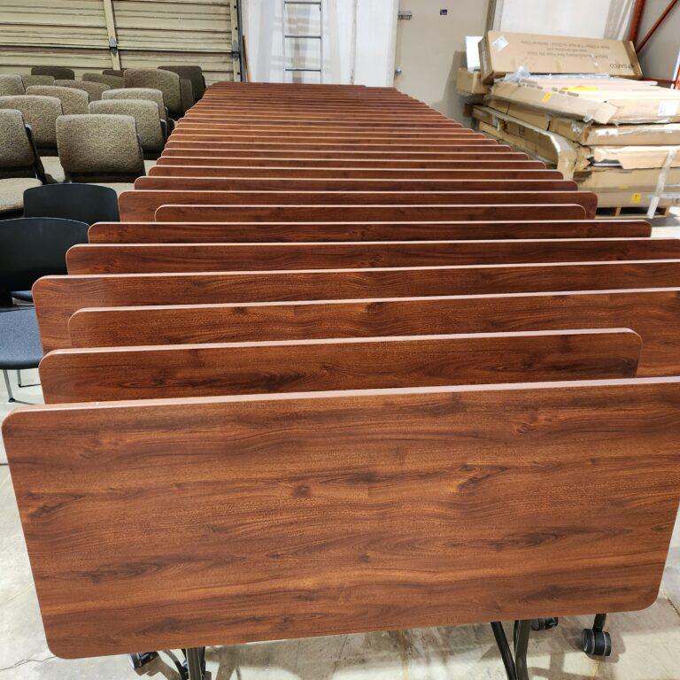 A row of nested training tables with wood-grain laminate tops and dark metal rolling bases in a warehouse setting.