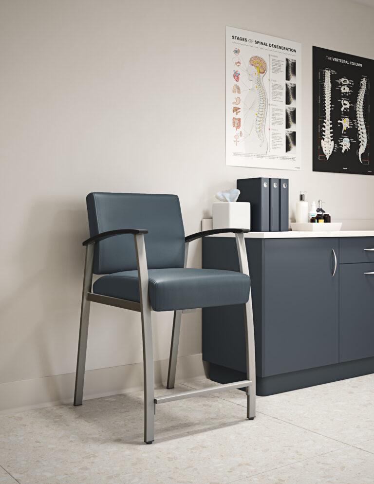 A Vitality stool with blue vinyl upholstery, a gray metal frame, and black armrests in a medical office setting next to a gray cabinet On the cabinet are a tissue box, 3 binders, and a collection of small bottles. Above the cabinet to the right of the stool are two posters - "STAGES OF SPINAL DEGENERATION" and "THE VERTEBRAL COLUMN."