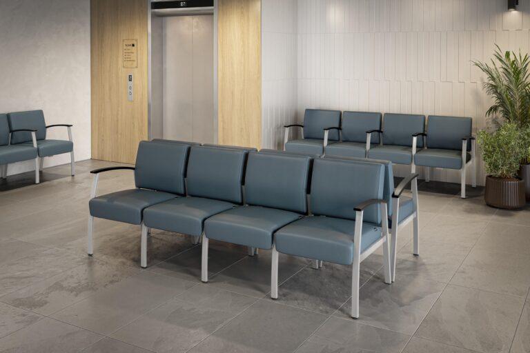 Multiple rows of Vitality beam seating with blue upholstered cushions, gray metal frames, and black armrests in an office waiting area featuring an elevator and potted plants.