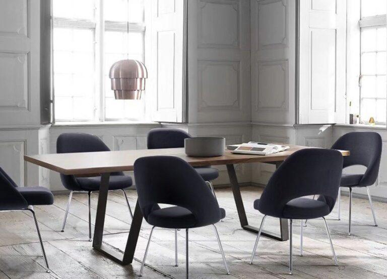 Several Melanie chairs with dark navy upholstery and chrome legs arranged around a wooden dining table in a bright room with white paneled walls.