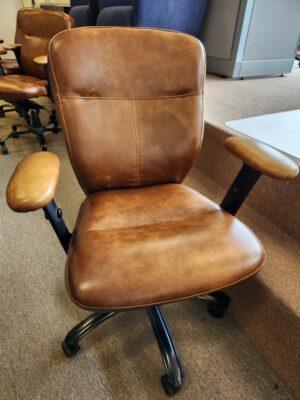 A mid-back office chair upholstered in cognac brown distressed leather or faux-leather with horizontal and vertical stitching details. The chair features padded armrests on black supports and a black five-star swivel base with casters, shown in an office showroom setting.