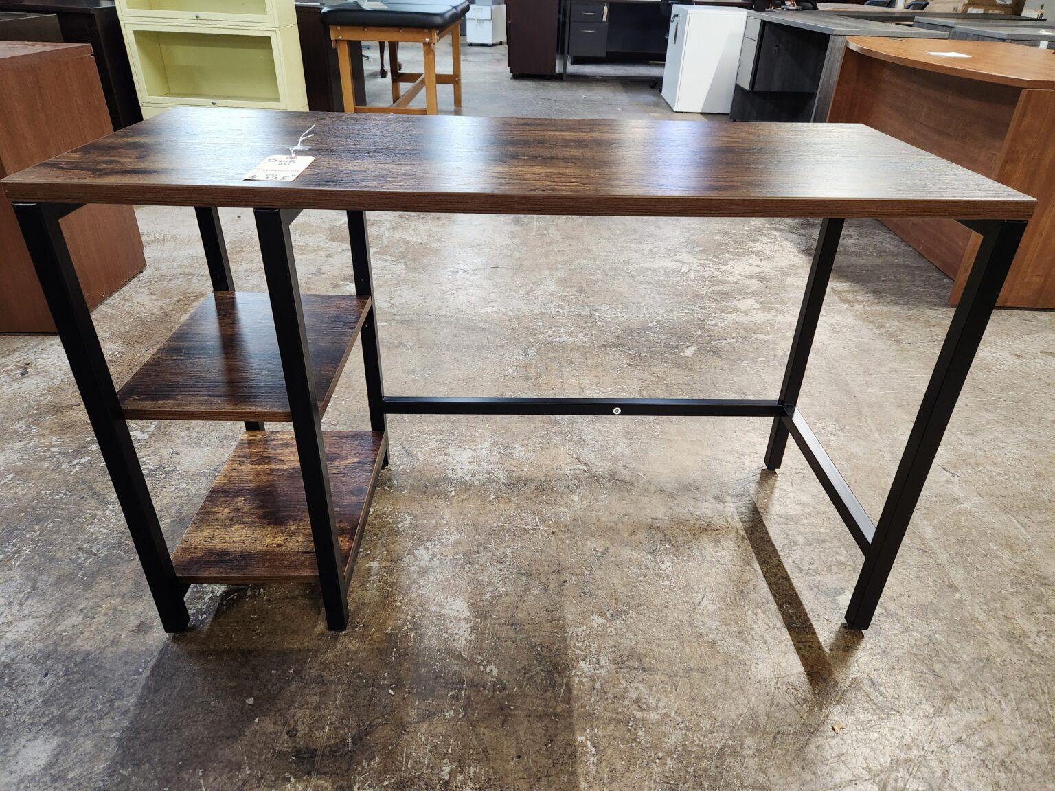 A rustic industrial computer desk featuring a dark walnut-finished wood grain tabletop and two integrated side storage shelves. The unit is supported by a minimalist matte black square steel frame with a cross-bar for added stability. The desk is positioned in a warehouse or showroom setting with concrete flooring and various other office furniture pieces visible in the background, including desks and shelving units.