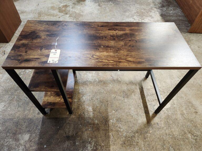 A high-angle view of a rustic industrial computer desk featuring a dark walnut-finished wood grain tabletop and two integrated open storage shelves. The desk is built with a minimalist matte black square steel frame and includes a price tag attached to the surface. The unit is shown in a warehouse or showroom setting with a concrete floor and portions of other office furniture visible in the surrounding area.
