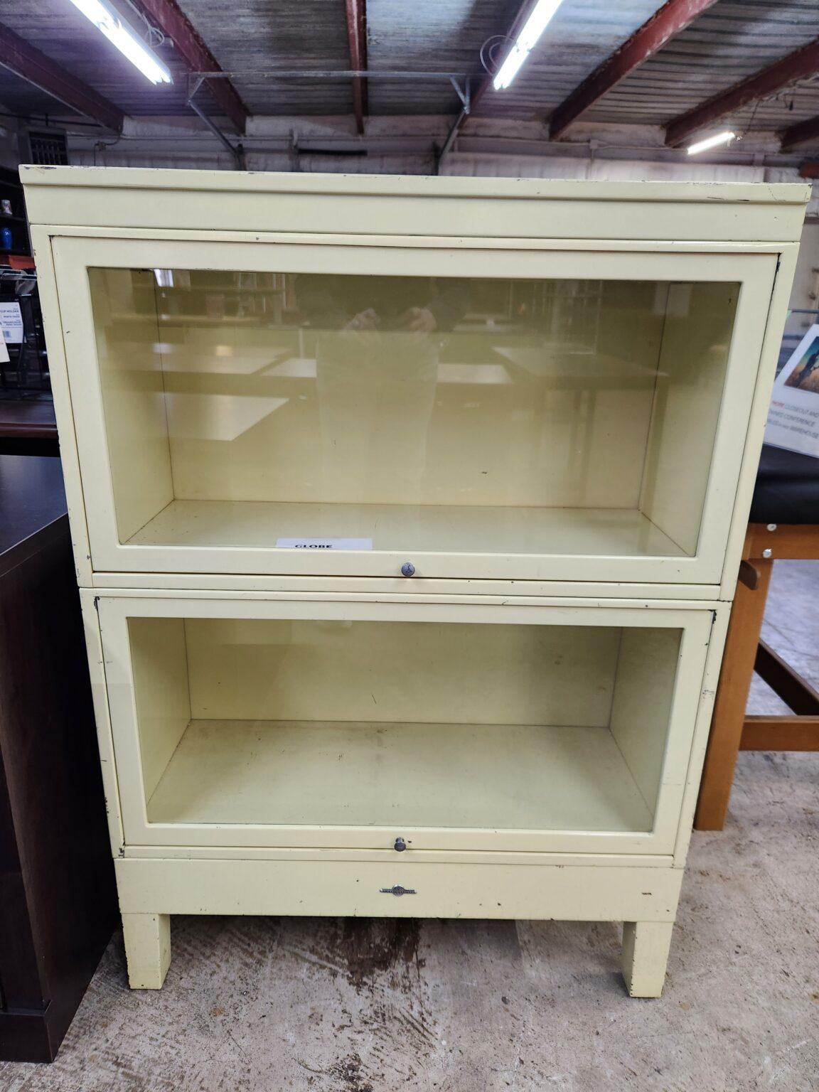 A vintage Globe-Wernicke barrister bookcase featuring a cream-colored metal frame with two glass-fronted stacking compartments. Each section includes a flip-up glass door with a small circular pull knob and a visible manufacturer's emblem on the base. The unit is shown in a warehouse setting with concrete floors, industrial overhead lighting, and other miscellaneous office furniture pieces in the background.