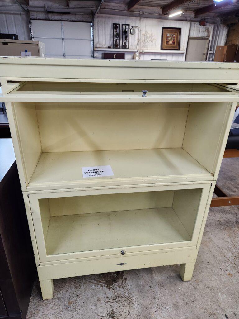 A vintage Globe-Wernicke barrister bookcase featuring a cream-colored metal frame with the top glass-fronted door shown in an open, recessed position. The unit consists of two stacking compartments, each equipped with flip-up glass doors and small circular pull knobs, with a descriptive label placed on the middle shelf. The bookcase is situated in an industrial warehouse environment featuring concrete floors, a large white garage door, and various pieces of furniture and wall decor in the background.