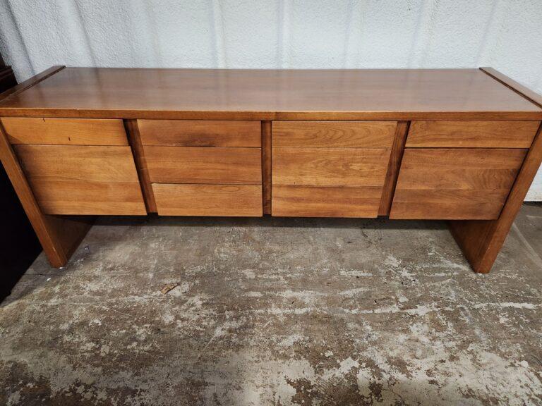 A mid-century modern style wood credenza in a warm brown finish with all drawers closed, featuring a rectangular top with raised side edges. The unit is divided into four vertical drawer sections and supported by thick slab-style legs on a concrete floor.