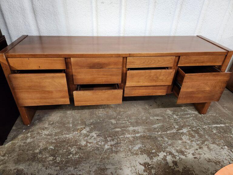 A mid-century modern style wood credenza in a warm brown finish, featuring four vertical sections of drawers with several drawers pulled open to show the interior storage. The unit has a flat rectangular top with raised side edges and sits on slab-style legs over a concrete floor against a white textured wall.