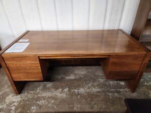 A mid-century modern style executive desk crafted from wood with a warm brown finish, featuring a wide rectangular top, two pedestals with built-in drawers, and a recessed modesty panel. The desk sits on a concrete floor against a white textured wall, with two white paper labels placed on the top left surface.