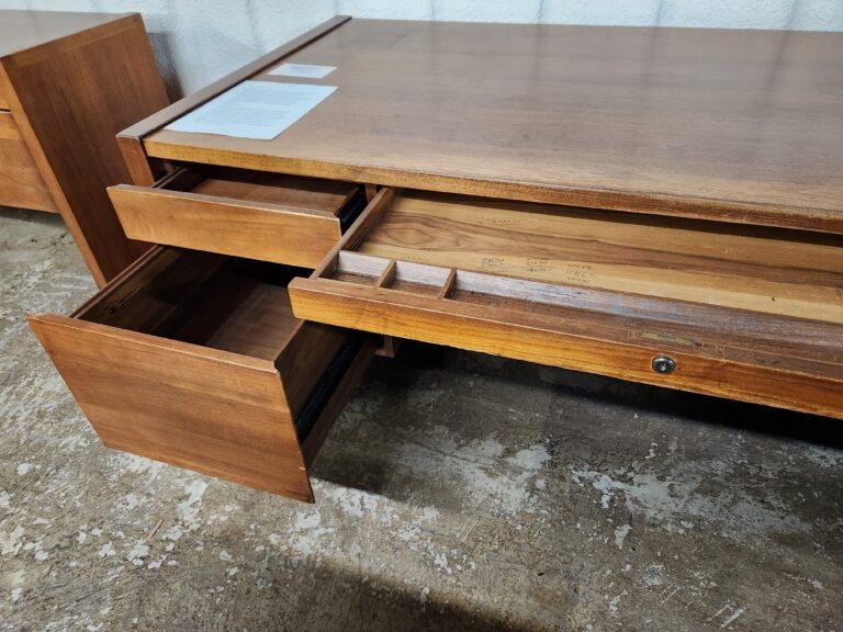 A mid-century modern style wood desk in a warm brown finish with two open side drawers and a pulled-out center pencil tray featuring built-in organizational compartments. The pencil tray includes handwritten markings on the wood interior, and the desk surface has two white paper labels on the top left corner.