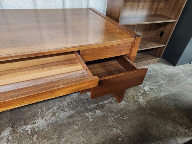 A wood executive desk in a warm brown finish featuring a pull-out center pencil tray and an open side drawer. The desk is positioned on a concrete floor next to a matching wood bookshelf with adjustable shelves.