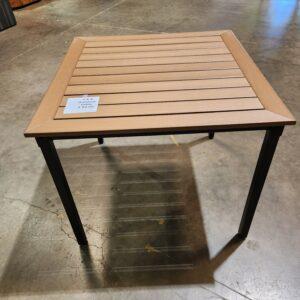 A square 4x4 outdoor table featuring a light wood-look slatted tabletop with a matching picture-frame border. The unit is supported by a minimalist black metal four-legged frame and includes a white descriptive price tag on the surface. The table is positioned on a cracked concrete floor within a warehouse or showroom setting, with its shadow clearly visible beneath the slatted design.