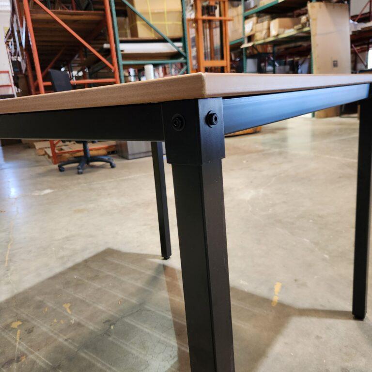 A low-angle, close-up view of a square outdoor table highlighting the structural assembly of the light wood-look tabletop and its black metal frame. The image focuses on the corner joint where the square metal leg is secured to the apron with visible hex-head bolts. The table is set in a warehouse environment featuring concrete floors and industrial pallet racking loaded with boxes in the background.