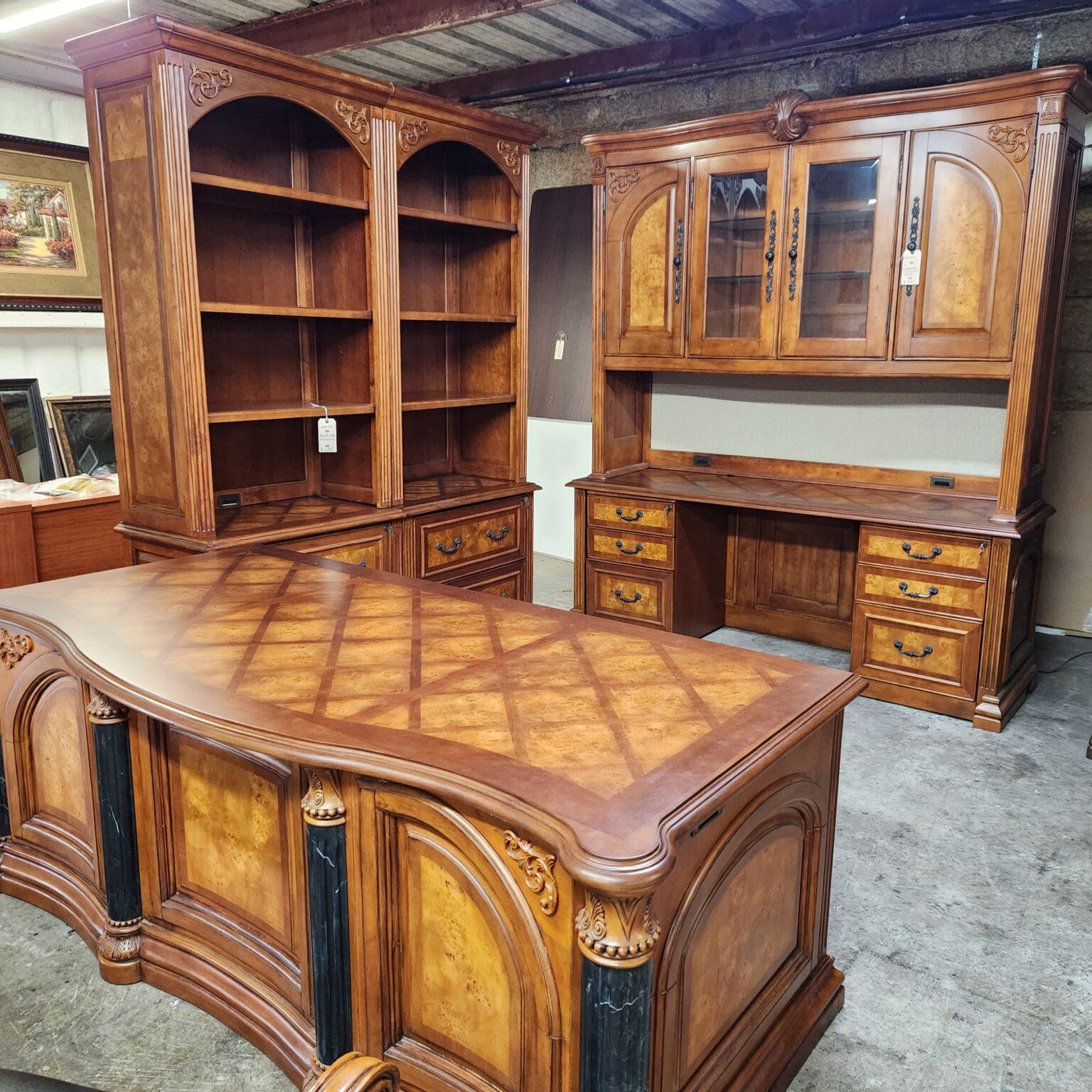 A dark wood executive office furniture suite in a warehouse setting, featuring a curved desk with dark fluted columns and a diamond-patterned top. The set also includes two joined open bookcases with arched tops and a matching credenza with a glass-front hutch, all detailed with lighter burl wood veneer accents, carved scrollwork, and metal drawer pulls.