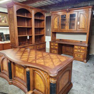 A dark wood executive office furniture suite in a warehouse setting, featuring a curved desk with dark fluted columns and a diamond-patterned top. The set also includes two joined open bookcases with arched tops and a matching credenza with a glass-front hutch, all detailed with lighter burl wood veneer accents, carved scrollwork, and metal drawer pulls.
