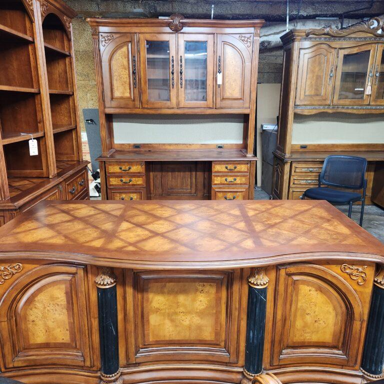 A dark wood credenza and hutch in a warehouse setting. The hutch features glass-front center doors and solid outer doors with arched burl wood veneer panels above a fabric tackboard, while the credenza base includes a diamond-patterned top, fluted column details, and six drawers with burl wood fronts and dark metal bail pulls.