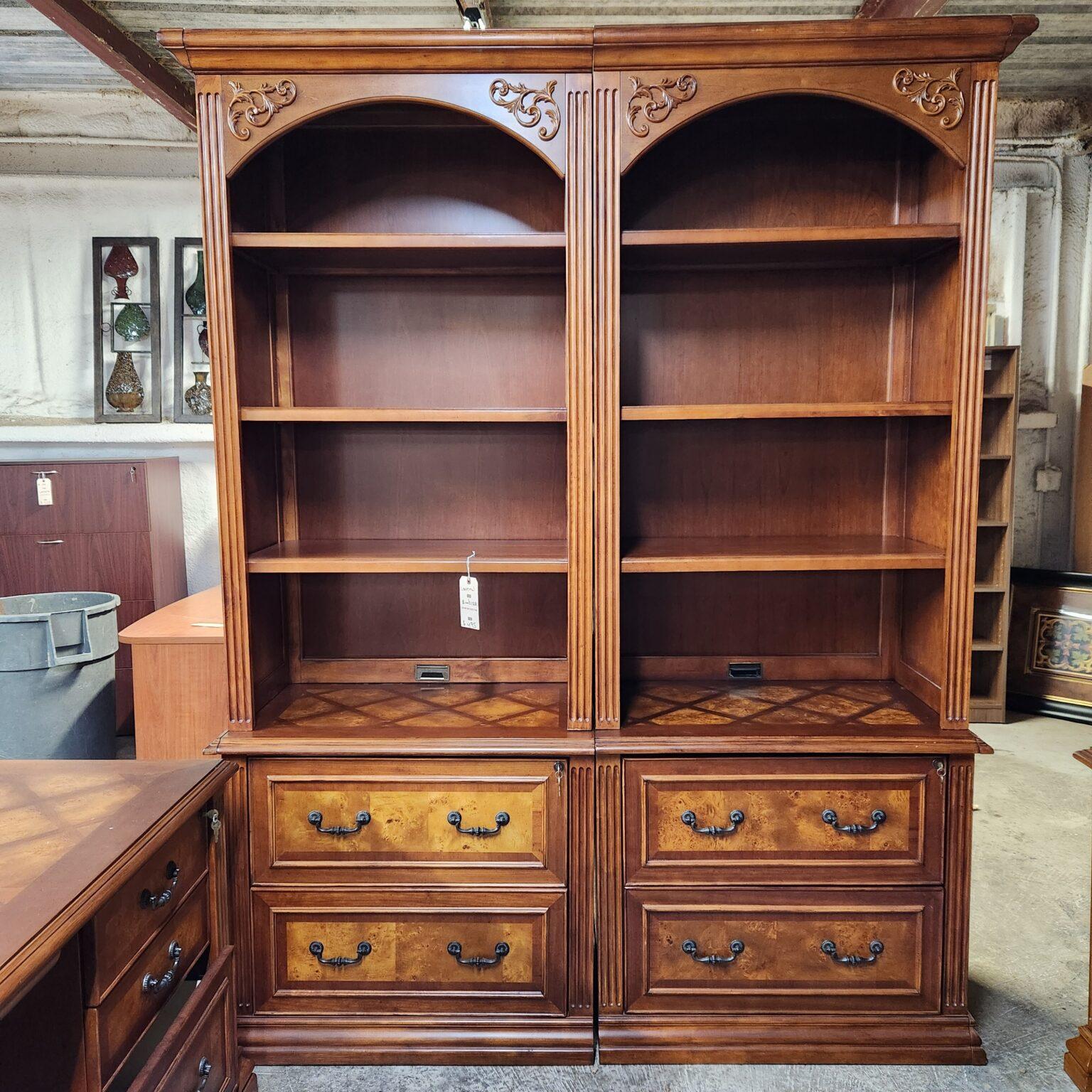 Two side-by-side dark wood bookcase cabinets in a warehouse setting. The upper sections feature open shelving with arched tops, carved scroll accents, and fluted columns, while the lower sections include diamond-patterned surfaces, wire grommets, and two large drawers with burl wood veneer fronts and metal bail pulls.