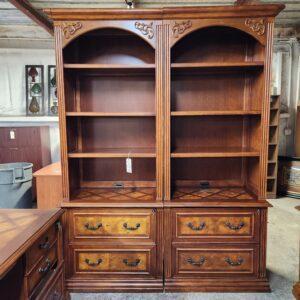 Two side-by-side dark wood bookcase cabinets in a warehouse setting. The upper sections feature open shelving with arched tops, carved scroll accents, and fluted columns, while the lower sections include diamond-patterned surfaces, wire grommets, and two large drawers with burl wood veneer fronts and metal bail pulls.