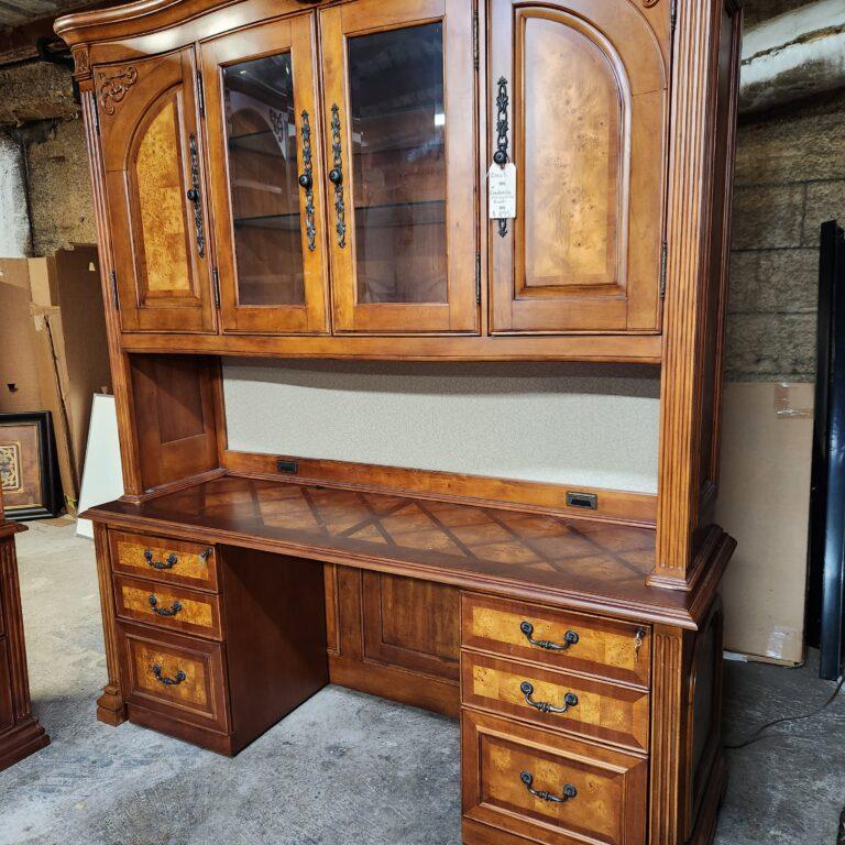 A dark wood credenza and hutch in a warehouse setting. The hutch features glass-front center doors and solid outer doors with arched burl wood veneer panels above a fabric tackboard, while the credenza base includes a diamond-patterned top, fluted column details, and six drawers with burl wood fronts and dark metal bail pulls.