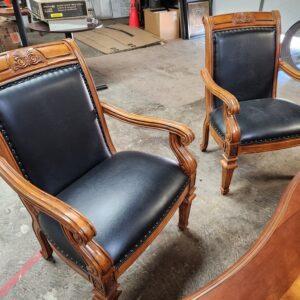 Two wooden armchairs in a warehouse setting, positioned near a curved wooden desk. The chairs feature medium-tone carved wood frames with scrolled armrests and fluted front legs, along with black upholstery accented by metal nailhead trim along the seats and backrests.